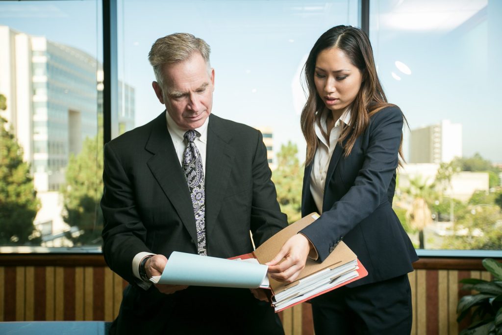 Derek Pakiz attorney in suit with woman paralegal holding papers in office in front of window blue sky green trees professional